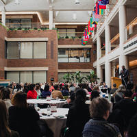 A wide shot of an atrium with an audience sitting at round tables listening to Harvey Charles speak