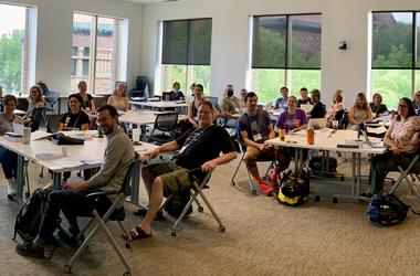 Members of CARLA sit in a classroom setting, with several people gathered at tables while sitting in chairs. There are large windows in the back of the room.